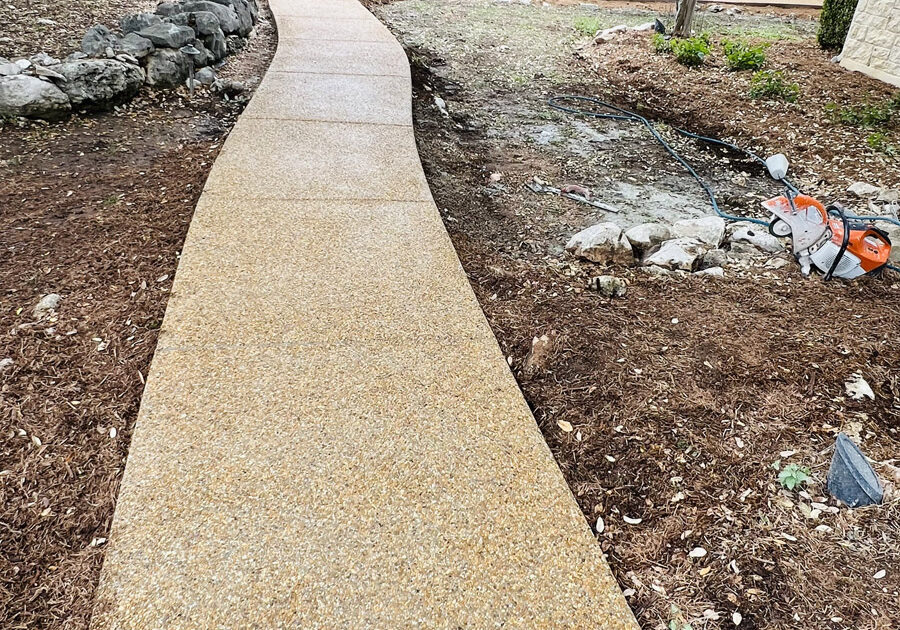 A newly laid beige gravel pathway curves gracefully through a park-like setting, leading towards a home with a stone facade. Landscaping efforts are evident with mulch beds, a rock wall, and a leaf blower resting on the side, suggesting ongoing maintenance.