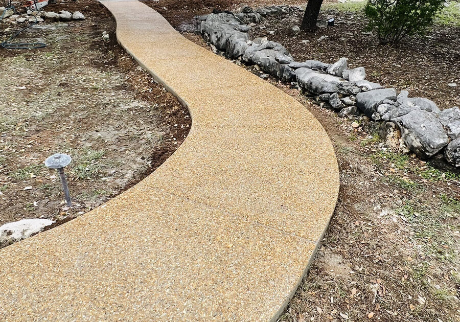 An elegantly winding pebble walkway in a San Antonio park, bordered by natural stone and interspersed with mature trees. The path leads towards the park’s entrance, with a glimpse of construction equipment in the distance.