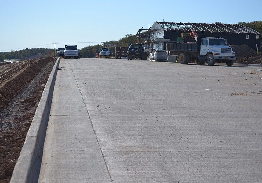 Newly constructed concrete roadway with vehicles parked along it and construction activity in the distance under a clear sky.