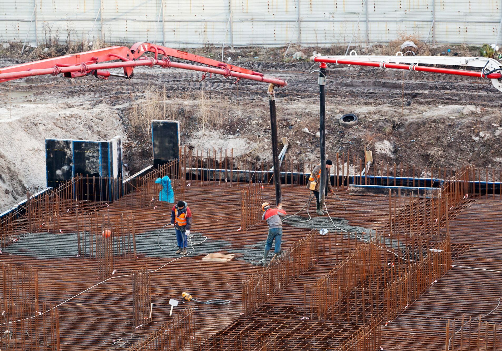 Workers pour concrete over rebar at construction site with red boom pump and excavation background.
