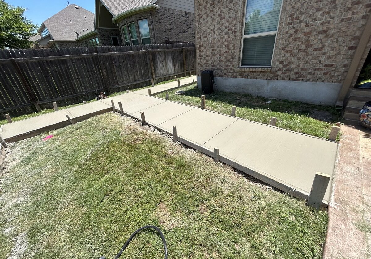 Freshly poured concrete walkway sections with wooden forms in a sunny backyard beside a brick house in San Antonio.