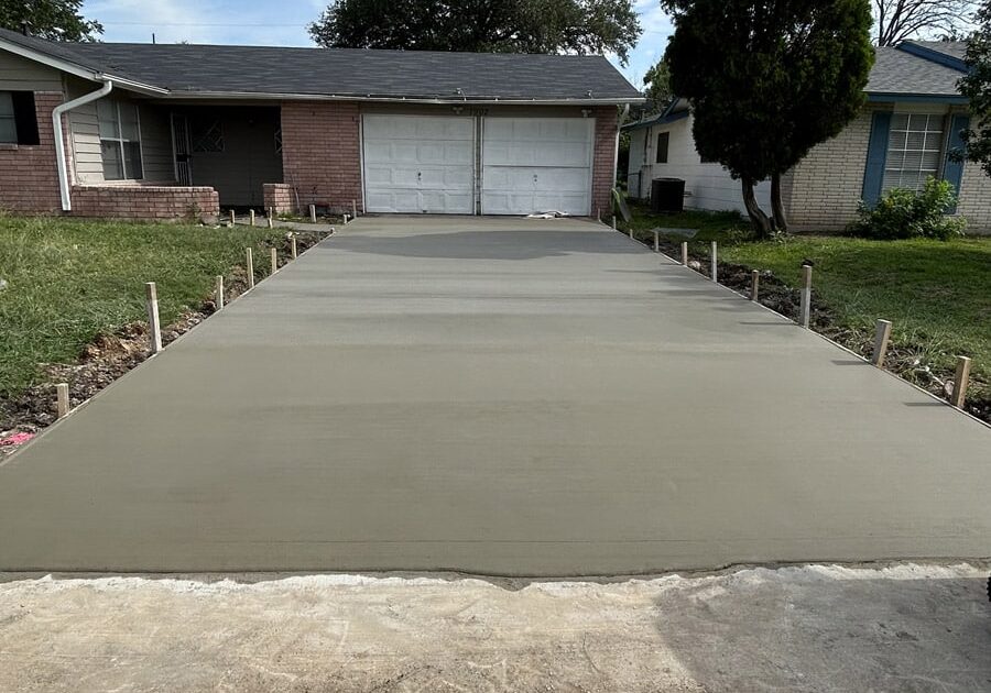 Residential driveway with fresh concrete and formwork, leading to a closed garage door, with a lawn and trees on either side.