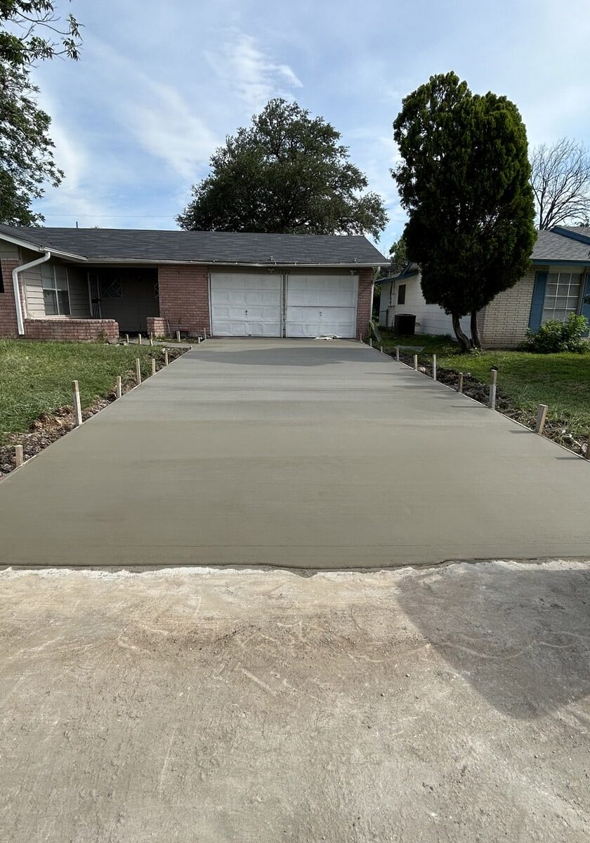 Residential driveway with fresh concrete and formwork, leading to a closed garage door, with a lawn and trees on either side.