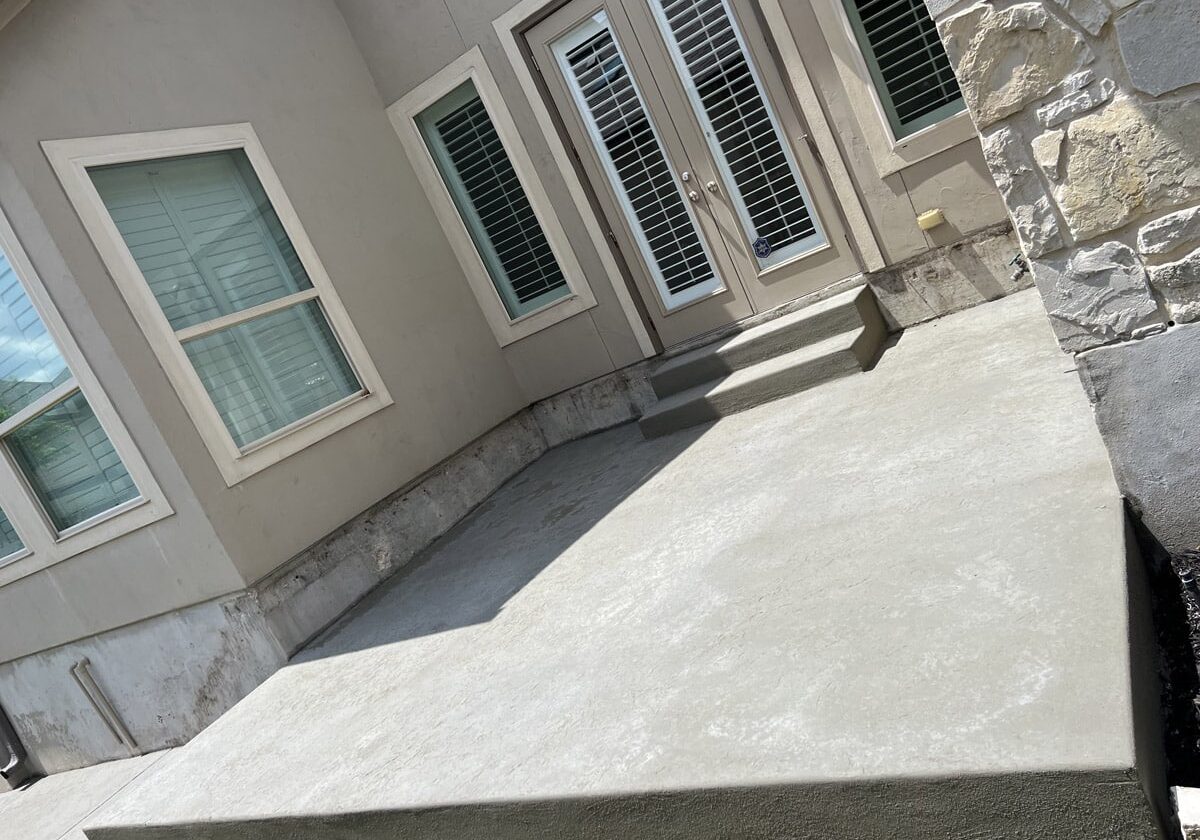 A freshly poured concrete porch in front of a house with a textured stucco facade and stone accents, under a bright sky in San Antonio.