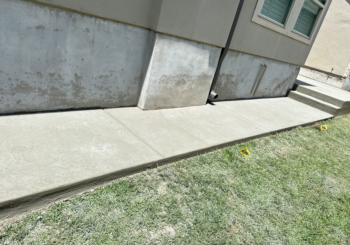 New concrete sidewalk installed against the exterior wall of a house, with fresh green grass on the side in San Antonio.