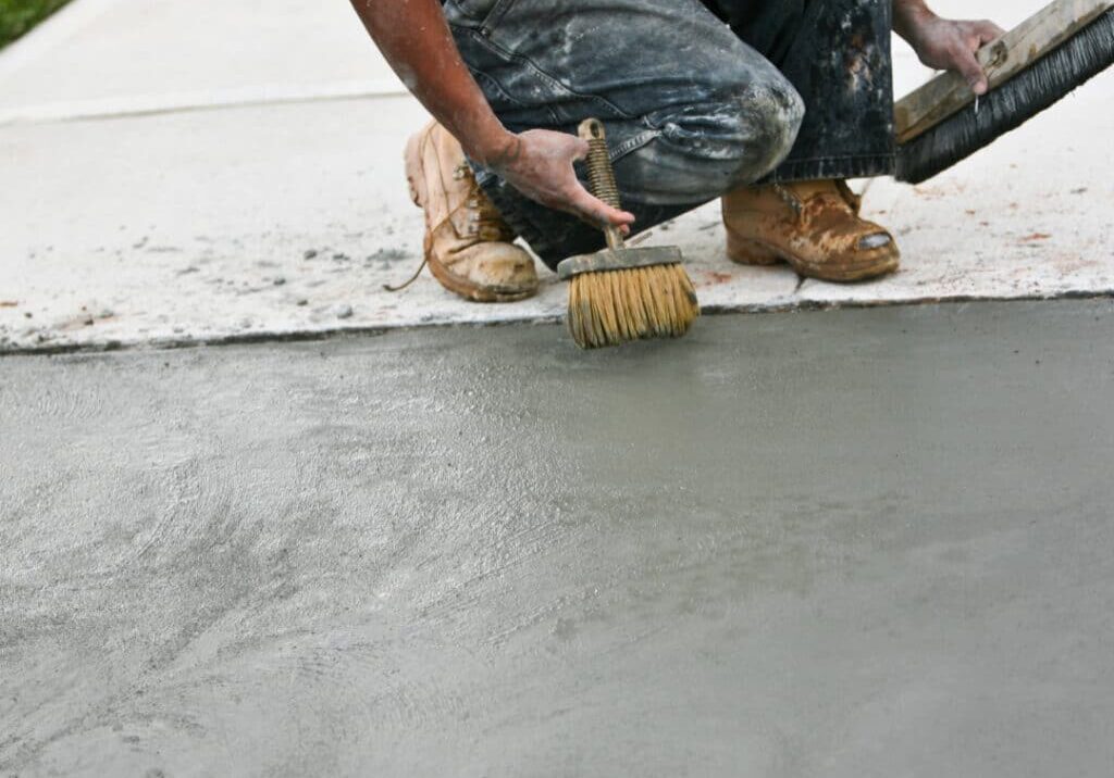 A construction worker crouched down applying a brush finish to fresh concrete on a sidewalk, wearing dirty work clothes and boots.